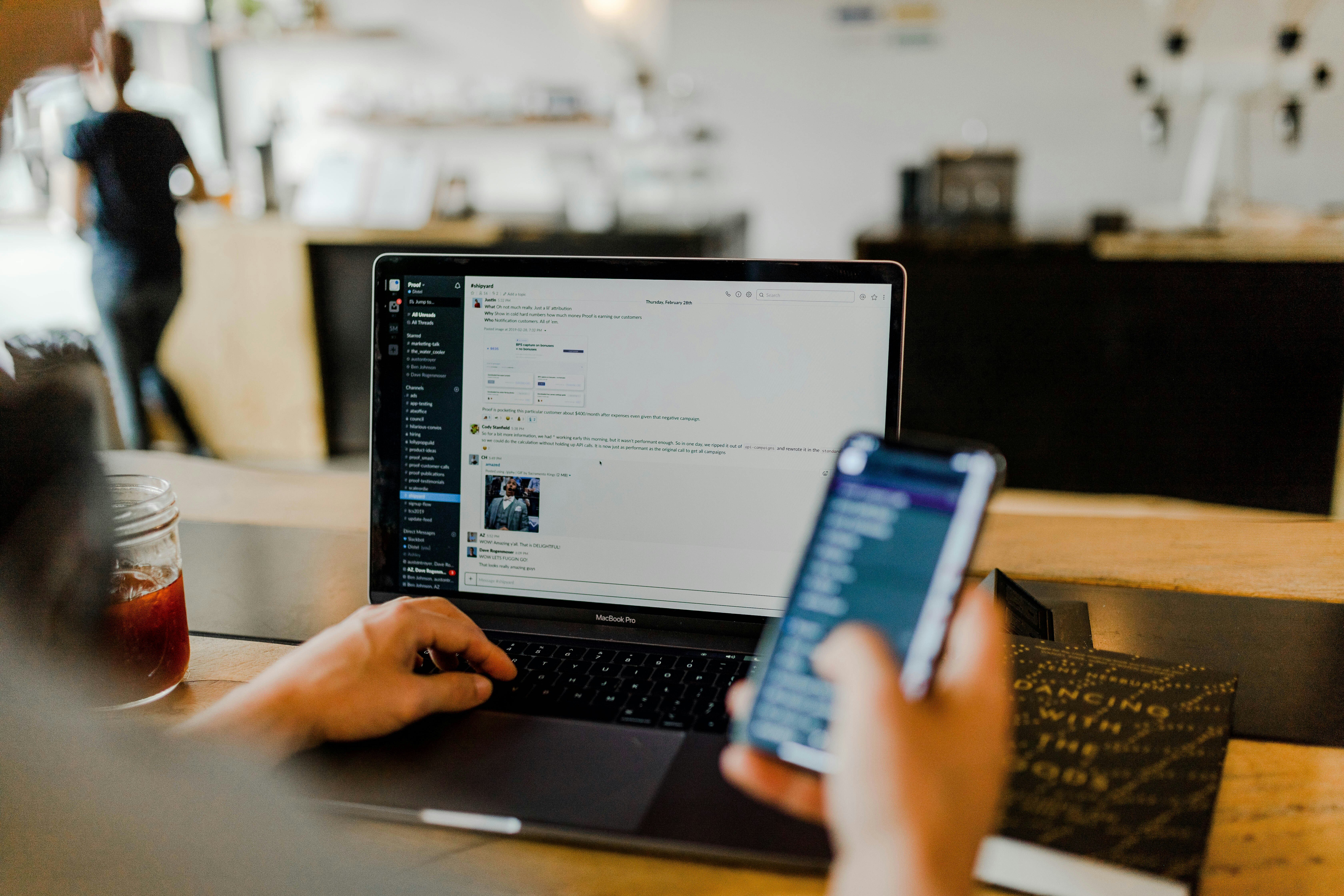 A person using a smartphone and laptop simultaneously in a casual workspace, with messaging apps open on both devices.