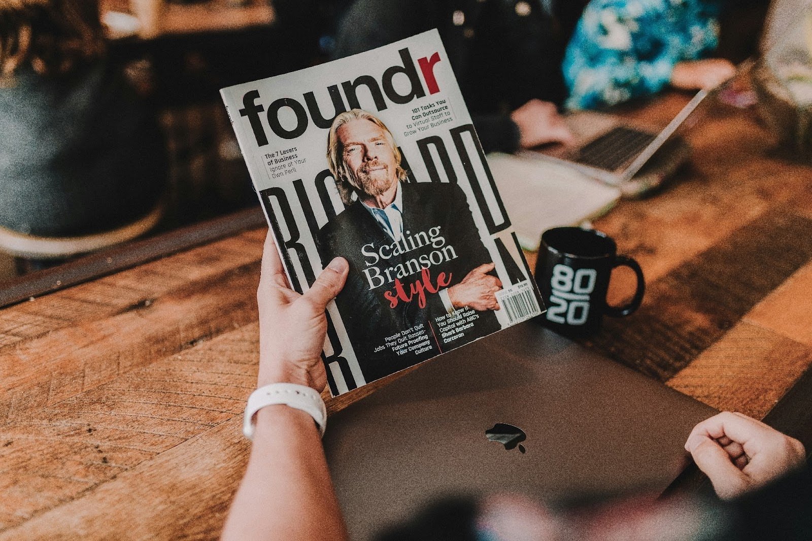 Person holding a Foundr magazine with Richard Branson on the cover, titled 'Scaling Branson Style,' with a black '80/20' mug and a MacBook on the table. The image reflects entrepreneurial leadership and business growth, as discussed on the WorkBuzz blog.