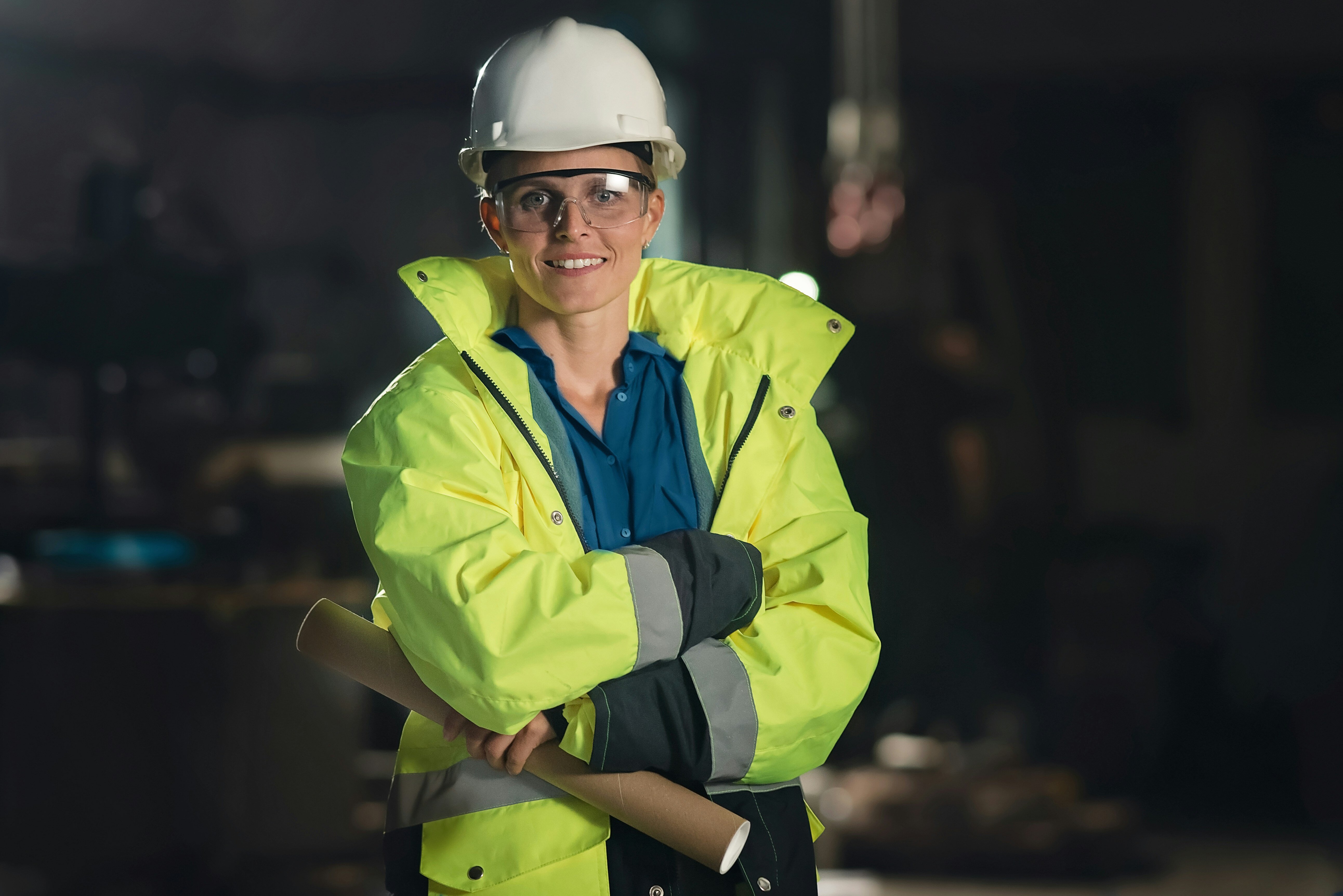 A female construction worker wearing a hard hat and high-visibility jacket smiling and holding a rolled-up blueprint, with a dimly lit industrial setting in the background.