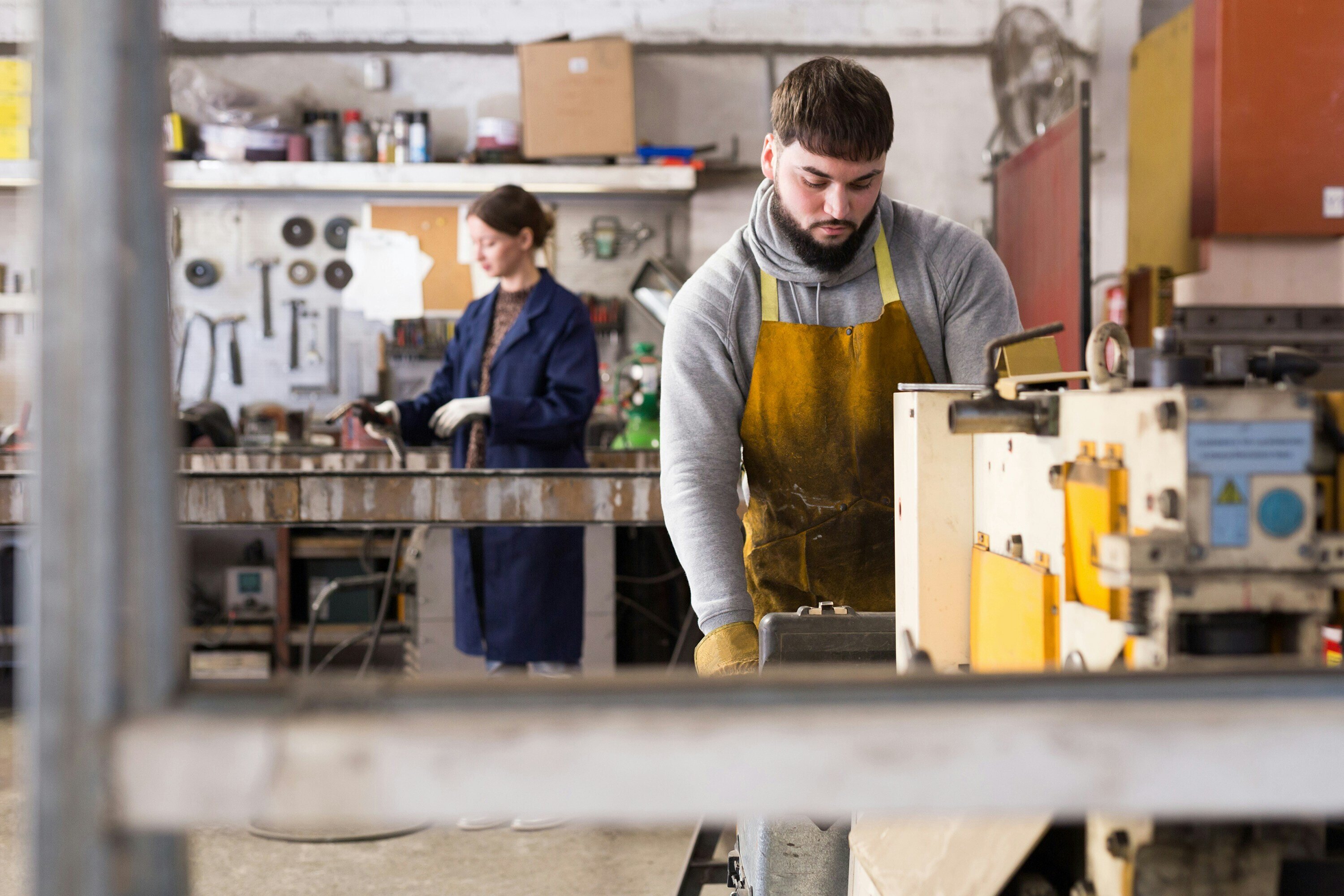 A man in a yellow apron working at his station in a workshop, with a female in a blue overcoat working at her station behind him, as seen on the WorkBuzz website.