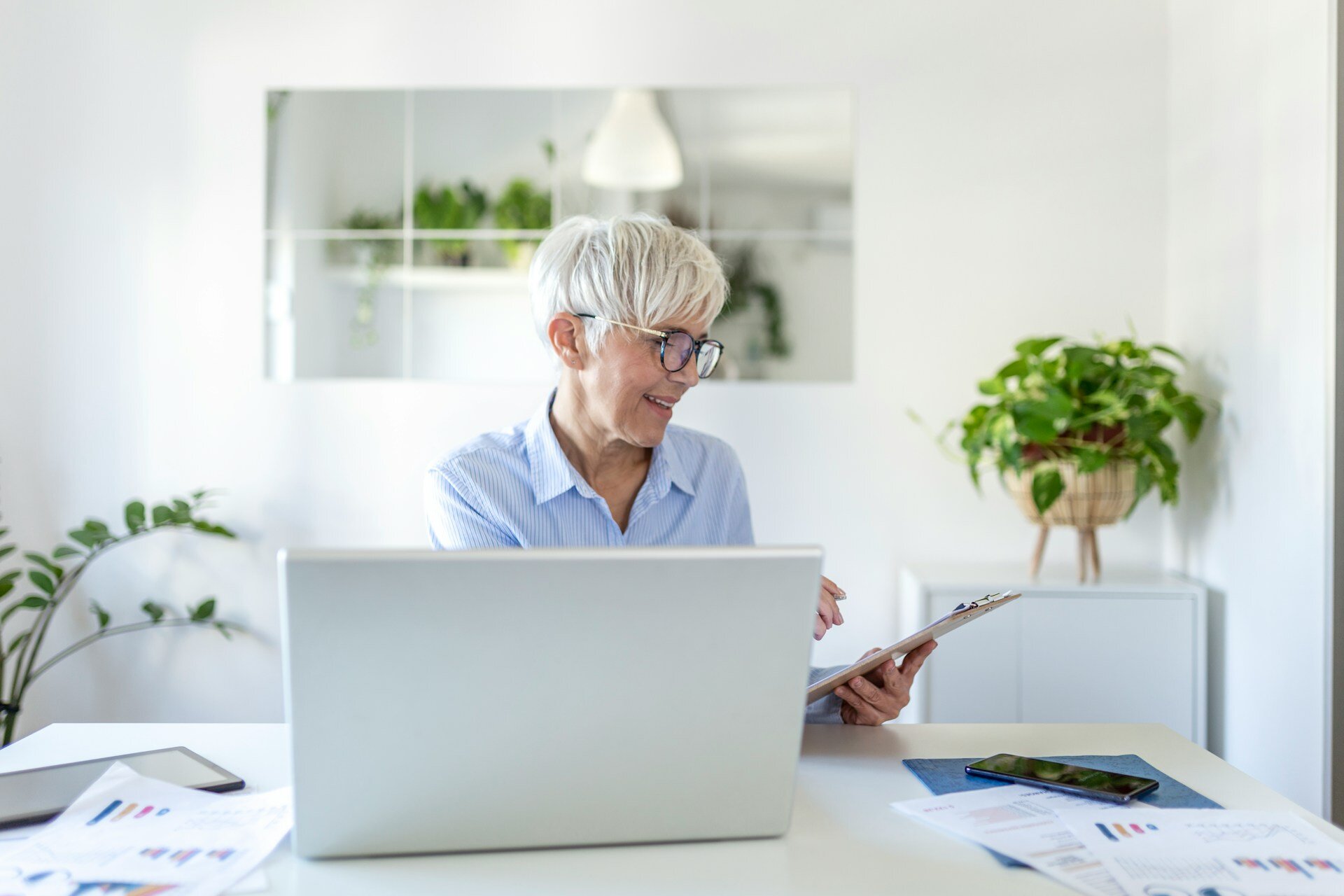 Smiling older woman with white hair works at a desk with a laptop and clipboard, surrounded by plants, as seen on the WorkBuzz website.