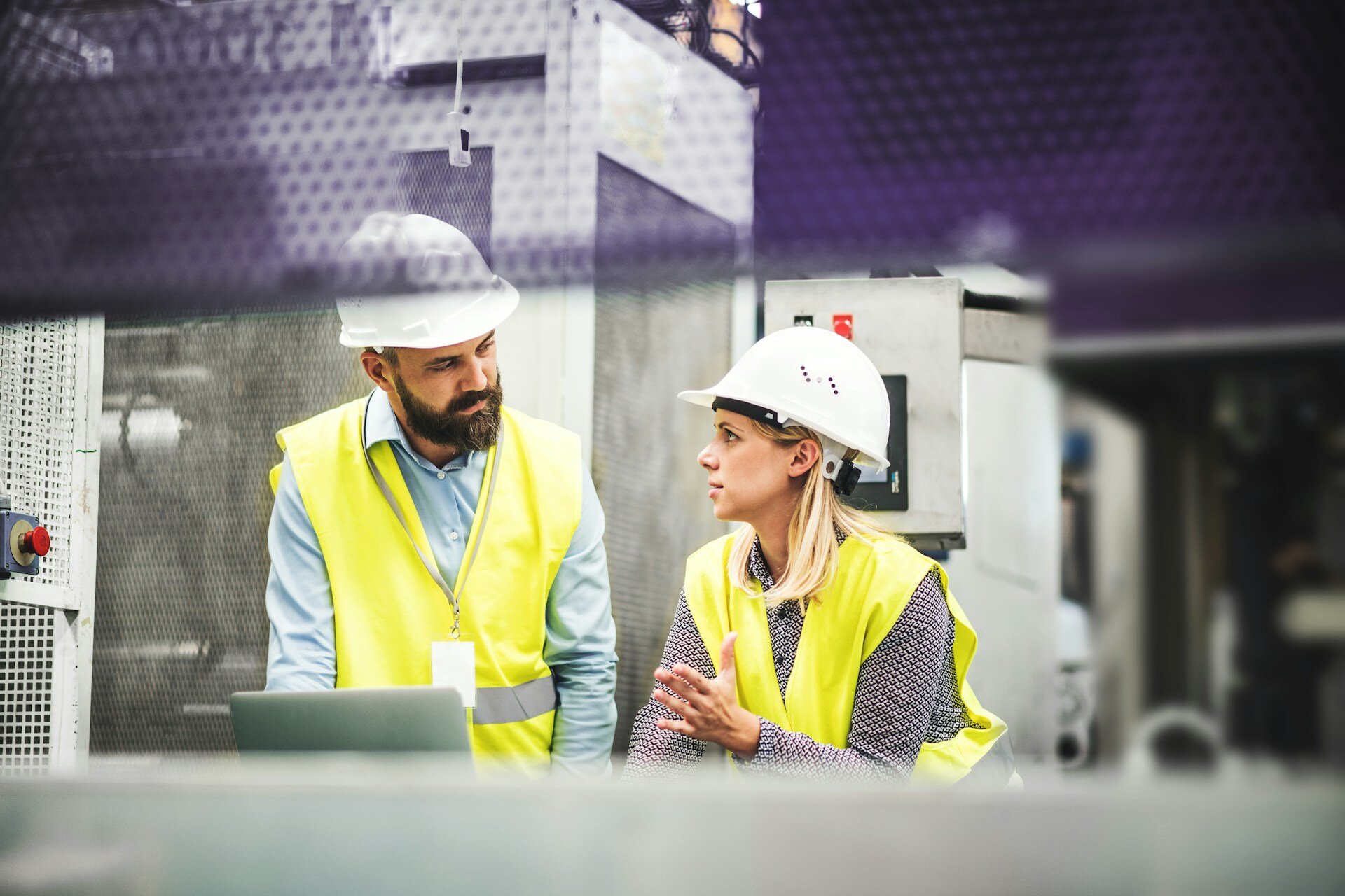 Man and woman in high-visibility jackets and white hard hats discussing work inside a manufacturing facility, as seen on the WorkBuzz website.
